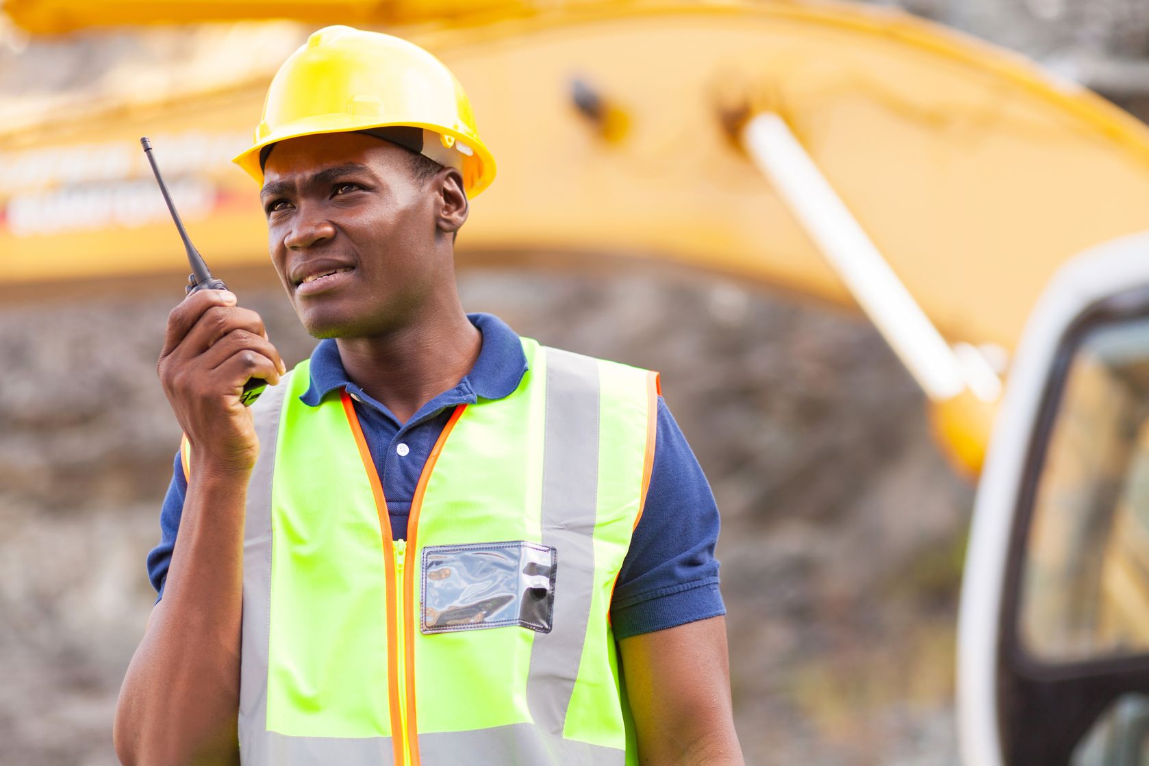 A construction worker in a hardhat and safety vest uses an RCA two-way radio while standing in front of an excavator.