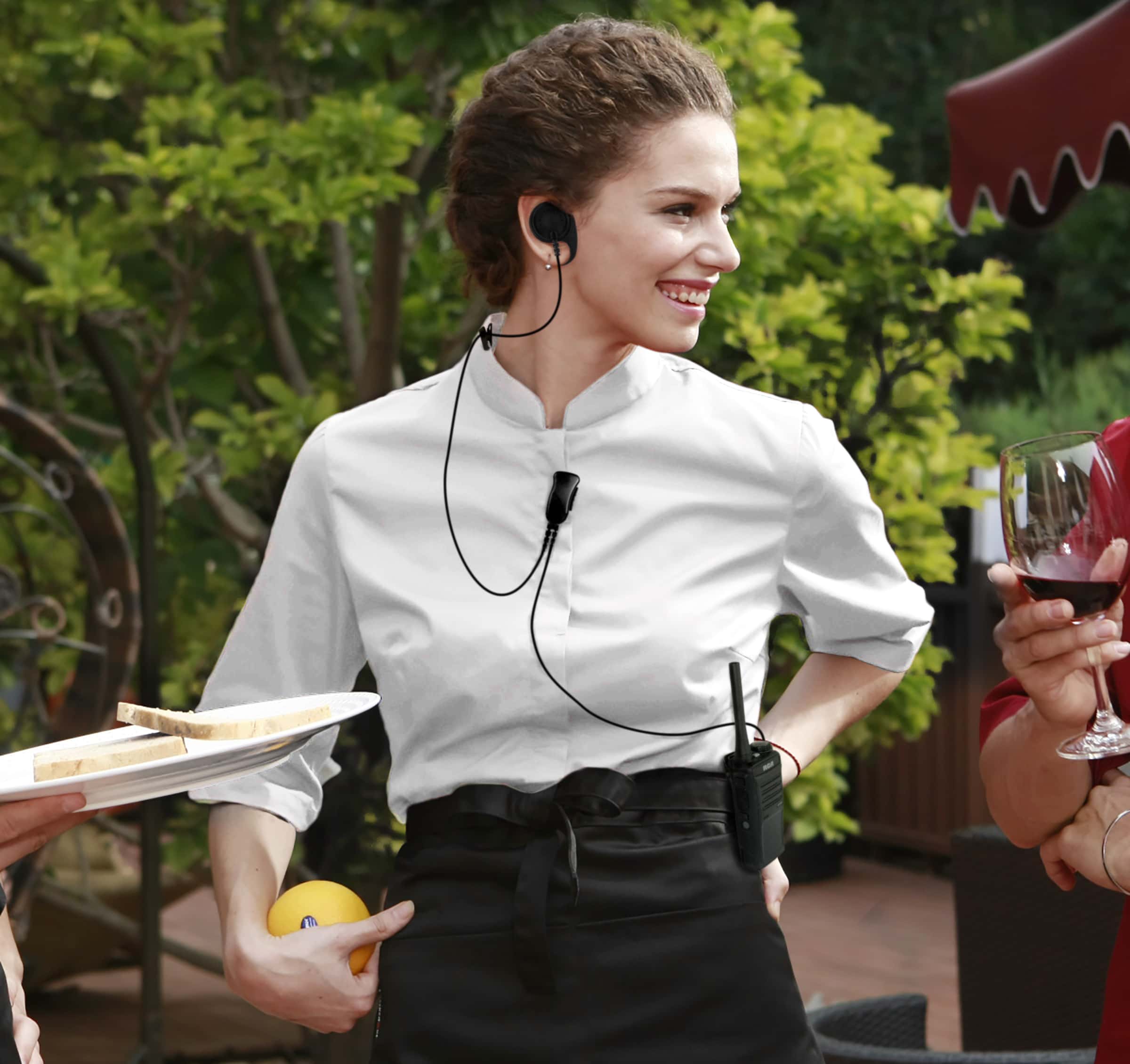 A restaurant server at a hotel smiles while talking with a customer outdoors. She uses a wired earpiece that connects with the RCA two-way radio clipped to her belt.