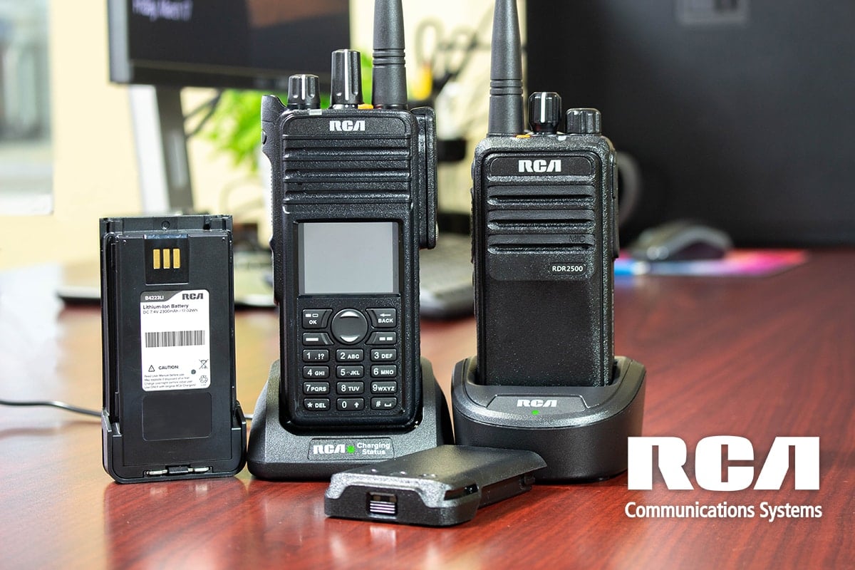 Two RCA two-way radio batteries are set beside two portable radios plugged into their charging cups on an office desk.